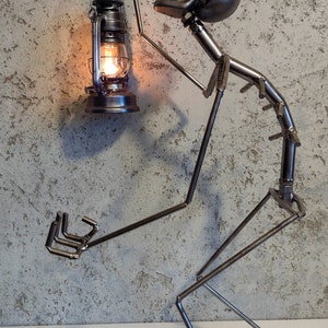 May include: A metal sculpture of a figure holding a lantern. The figure is made of welded metal rods and has a skull-shaped head. The lantern is lit with a warm, orange glow. The sculpture is on a white surface.