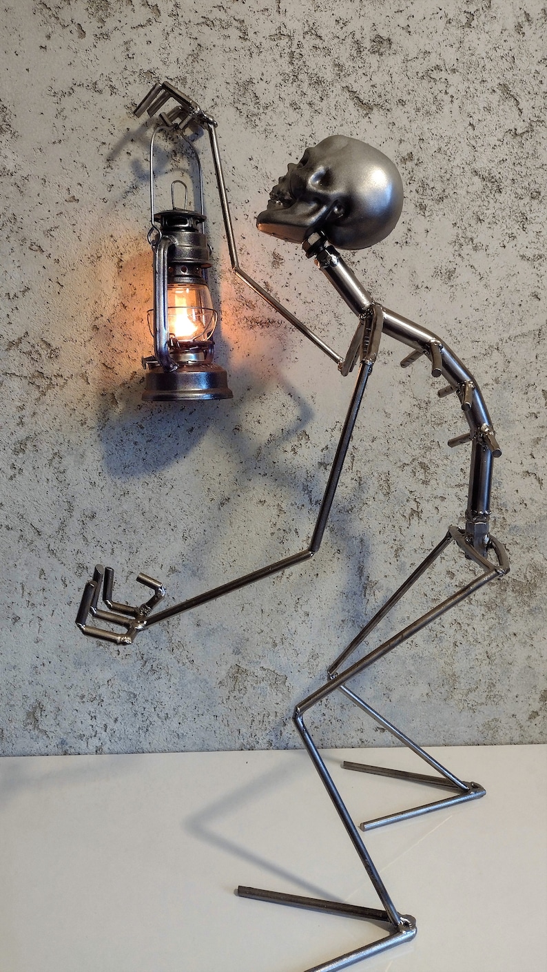 May include: A metal sculpture of a figure holding a lantern. The figure is made of welded metal rods and has a skull-shaped head. The lantern is lit with a warm, orange glow. The sculpture is on a white surface.