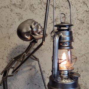 May include: A metal sculpture of a skeleton holding a lantern with a lit flame. The skeleton is silver and the lantern is dark gray. The background is a textured, light gray wall.