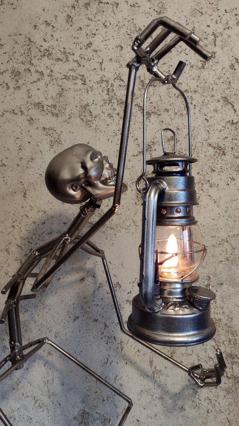 May include: A metal sculpture of a skeleton holding a lantern with a lit flame. The skeleton is silver and the lantern is dark gray. The background is a textured, light gray wall.