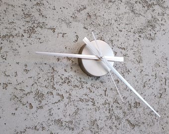 Puede incluir: Un reloj de pared moderno con una esfera de metal cepillado y manecillas blancas minimalistas. El reloj está montado en una pared texturizada de color gris claro. La aguja horaria mide unos 25 cm.