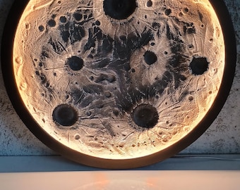 Puede incluir: Una obra de arte mural redonda e iluminada que representa la superficie de la luna. La obra presenta una superficie texturizada y con cráteres en tonos grises y marrones, con una luz cálida y brillante que emana del interior. La pieza está enmarcada por un borde circular oscuro.