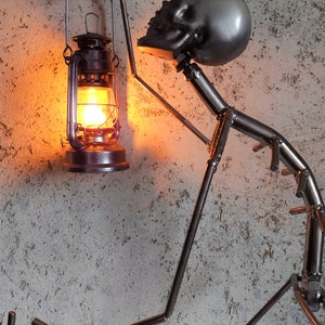 May include: A metal sculpture of a figure holding a lit lantern. The figure has a skull-shaped head and is made of welded metal rods. The lantern is a traditional style with a glass globe and a warm, orange glow.