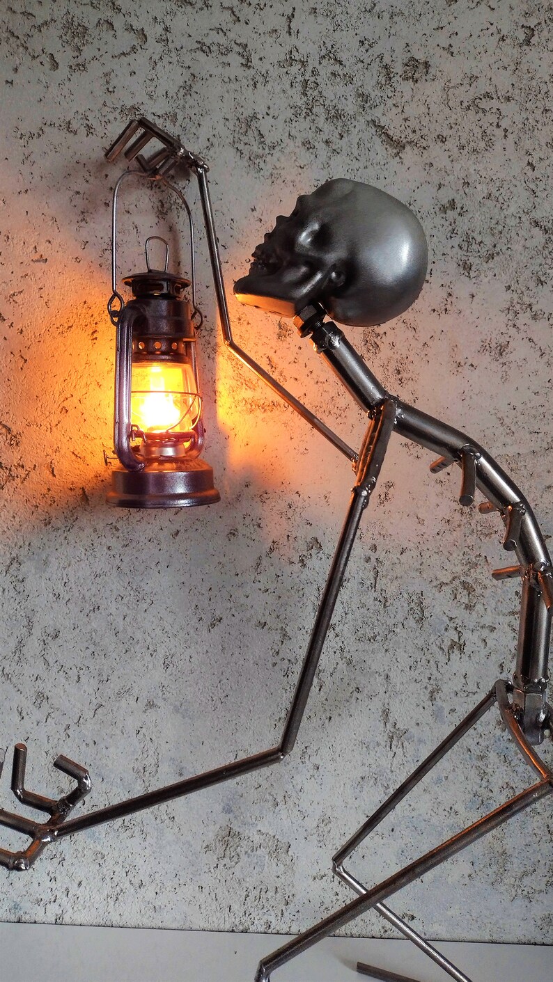 May include: A metal sculpture of a figure holding a lit lantern. The figure has a skull-shaped head and is made of welded metal rods. The lantern is a traditional style with a glass globe and a warm, orange glow.