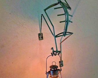 May include: A metal skeleton sculpture holding a vintage oil lamp. The skeleton is made of metal rods and has a silver skull. The lamp is black with a glass globe and is lit.
