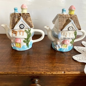 Könnte beinhalten: Zwei kleine Keramik-Teekannen in Form von Häusern mit einem Zifferblatt und einem Hasen auf der Vorderseite. Die Teekanne rechts trägt die Aufschrift "Mom Tea House".