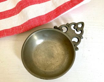 Peut inclure: Un petit bol en étain antique avec une poignée ornée. Le bol est rond et présente un aspect légèrement vieilli. Un tissu à rayures rouges et blanches est en arrière-plan, suggérant une esthétique rustique ou vintage.