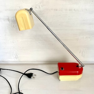 Könnte beinhalten: Eine Schreibtischlampe mit einem cremefarbenen, rechteckigen Lampenschirm und einem passenden cremefarbenen Sockel, der mit einer glänzenden roten Abdeckung versehen ist. Die Lampe hat einen langen, verstellbaren, verchromten Metallarm und ein schwarzes Kabel.