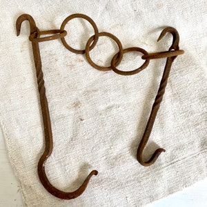 Pode incluir: Um gancho de metal enferrujado com uma alça torcida e uma série de anéis conectados. O gancho é feito de metal marrom escuro e tem uma aparência rústica.