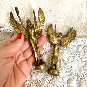 Peut inclure: Deux figurines d'anges dorées aux ailes déployées. Les anges sont posés sur une surface en dentelle blanche.