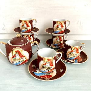 Op de afbeelding: Een vintage Japanse theeset met een theepot, kopjes en schotels. De set is diep bruin met gouden details en gedetailleerde illustraties van een figuur in traditionele kledij. De kopjes en schotels zijn op elkaar gestapeld.
