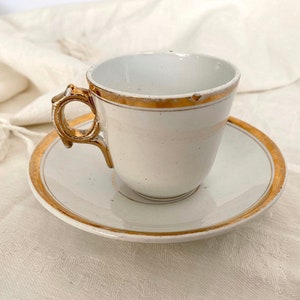 Könnte beinhalten: Eine weiße Porzellan-Teetasse und -Untertasse mit goldfarbenem Rand. Die Tasse hat einen einzigartigen, verzierten Henkel.