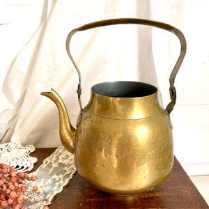 Può includere: Una teiera in ottone vintage con corpo arrotondato e manico curvo. La teiera ha una finitura usurata e antica, perfetta per aggiungere un tocco di fascino rustico alla tua cucina.