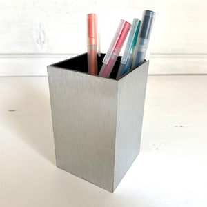 Può includere: Un portapenne in metallo argentato con finitura spazzolata. Il portapenne è pieno di diverse penne, tra cui una penna rossa, una penna blu e una penna verde.