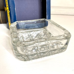 Op de afbeelding: Een vierkante asbak van helder glas met een diamantpatroon. De asbak staat op een wit oppervlak.