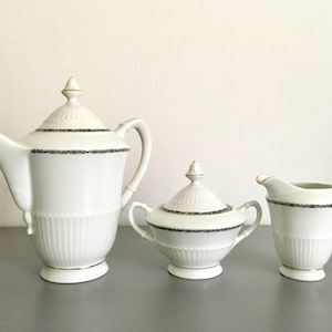 Pode incluir: Um conjunto de cafeteira, jarra de creme e açucareiro de porcelana branca com detalhes dourados e uma faixa com padrão preto e branco ao redor da parte superior de cada peça.