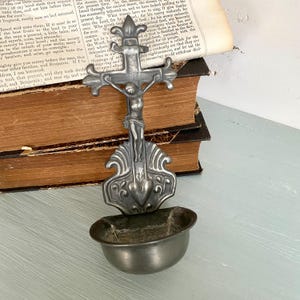 Könnte beinhalten: Ein Vintage-Weihwasserbecken aus Metall mit einem Kruzifix und einem kleinen Becken. Das Kruzifix zeigt Jesus Christus. Das Becken hat ein verziertes Design und ist wahrscheinlich aus Zinn oder einem ähnlichen Metall gefertigt. Es steht vor einem offenen Buch.