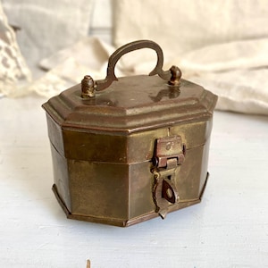 Könnte beinhalten: Eine kleine, achteckige Messingdose mit einem Scharnierdeckel und einem Schloss. Die Dose hat einen Griff oben und ist mit einem einfachen Design verziert.
