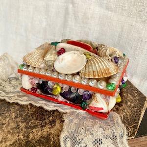Könnte beinhalten: Eine kleine, quadratische Schachtel, die mit Muscheln bedeckt ist. Die Schachtel ist aus Holz und rot lackiert. Die Oberseite der Schachtel ist mit einer Vielzahl von Muscheln bedeckt, darunter weiße, braune und rosa Muscheln. Die Schachtel ist mit kleinen, bunten Perlen verziert.