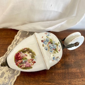 May include: White ceramic pulley with a floral design and a scene of women in a garden. The pulley has a metal hook and a white ceramic wheel.
