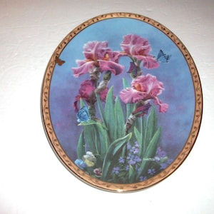May include: An oval decorative plate featuring a central illustration of pink iris flowers and green foliage against a blue background. Two small blue butterflies are visible among the blooms. The plate has a gold-toned border with a brown vine pattern.