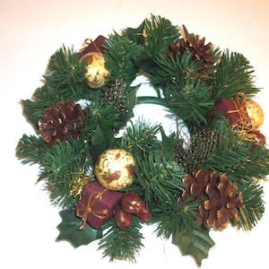 May include: A festive Christmas wreath with green faux pine needles, adorned with gold ornaments, burgundy ribbons, and brown pinecones. The wreath also features green holly leaves and small, decorative gift boxes.