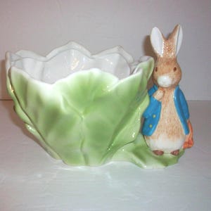 May include: A ceramic bowl shaped like a green cabbage leaf with a white interior. Attached to the side of the bowl is a standing brown rabbit figure wearing a blue jacket, with white fur on its chest and face, and holding an orange carrot.