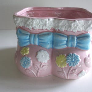 May include: A pink ceramic planter shaped like a baby bootie. It features a light blue bow, white floral trim, and embossed flowers in blue, white, and yellow. The planter has a square opening.