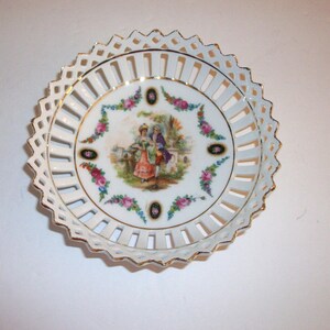 May include: White porcelain decorative plate with a pierced rim and gold trim. The center features a painted scene of a couple in period clothing, surrounded by floral accents. A vintage-style serving dish.