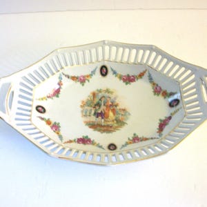 May include: An oval, white porcelain serving dish with a pierced rim and handles. The center features a painted scene of a couple in a garden, surrounded by floral and gold accents. The dish is approximately 12 inches long.