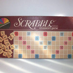 May include: A vintage Scrabble board game box with the game board visible. The box is maroon with the word "SCRABBLE" in white script. The game board is beige with colored squares and letter tiles scattered around.