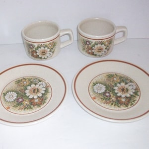May include: A set of two cream-colored coffee cups and saucers. Each piece features a floral design with white, yellow, and brown flowers. The rims of the cups and saucers have a brown border. The set is ideal for serving coffee or tea.