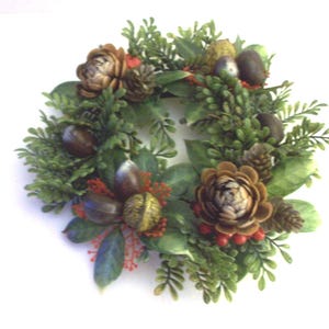 May include: A circular wreath featuring a variety of natural elements. The wreath includes green foliage, brown pinecones, and decorative accents. Red berries and acorn-like details add color and texture to the design.