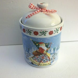 May include: A ceramic Christmas cookie jar with a white lid and a red and white checkered bow. The jar features a winter scene with Santa Claus carrying a Christmas tree. The rim is decorated with red flowers and green leaves.