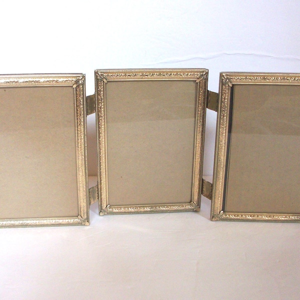 May include: A triptych photo frame featuring three rectangular panels connected by metal hinges. Each frame has a metallic gold finish with a textured, ornate border pattern and clear glass inserts, designed to hold three separate photographs.