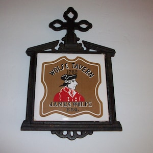 May include: A vintage Wolfe Tavern sign, featuring a portrait of James Wolfe, Esq. The sign, dated 1762, has a brown and white tile design with a black decorative frame and top cross. A classic piece of pub decor.