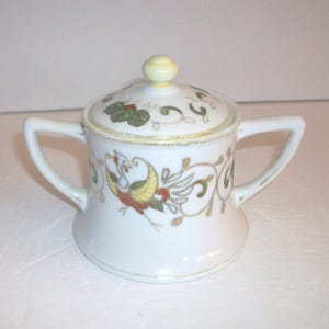 May include: White ceramic sugar bowl with a lid and two handles. The bowl is decorated with a colorful bird and floral design. The lid has a yellow knob. The rim and handles are trimmed with gold.