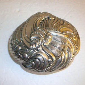 May include: A silver-colored, ornate, clam-shaped decorative box. The box features detailed, swirling patterns and textures across its surface. The design includes a combination of smooth curves and intricate beaded accents, creating a vintage aesthetic.