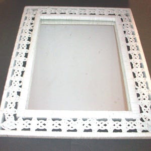 May include: A rectangular mirror featuring a white frame constructed from woven wicker material. The frame displays an intricate, repeating lattice pattern along its border, surrounding a clear, reflective glass center surface.