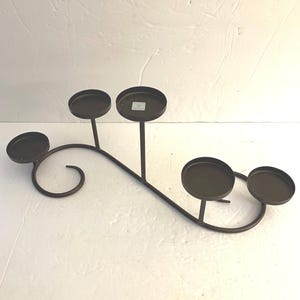 May include: A bronze-colored metal candle holder with five circular platforms for candles. The holder has a curved, flowing design with a decorative swirl at one end. The platforms are supported by vertical stems.