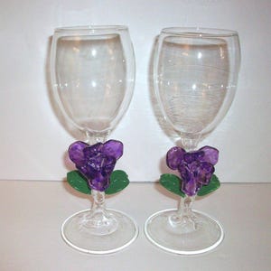 May include: Two clear wine glasses with a purple flower and green leaf design on the stem. The glasses have a rounded bowl and a clear base. The flower is a deep purple color.