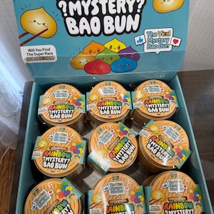 May include: A retail display box containing multiple round containers of Rainbow Mystery Bao Bun toys. The packaging is light blue with colorful illustrations of smiling bao buns and text that reads Rainbow Mystery Bao Bun throughout.