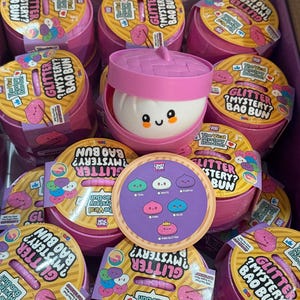 Könnte beinhalten: Eine Sammlung rosa Kunststoffbehälter in Form von Bambusdämpfern, beschriftet mit Glitter Mystery Bao Bun. Ein Behälter ist geöffnet und zeigt ein weißes, rundes, weiches Spielzeug mit einem lächelnden Gesicht. Eine kreisförmige Anleitung zeigt sechs Bao-Farben.