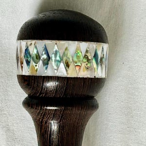 Może przedstawiać: Ciemnobrązowa drewniana laska z ozdobnym uchwytem. Uchwyt posiada pasmo iryzującej muszli abalonu w rombowy wzór, osadzone pomiędzy białymi segmentami. Złoty pas akcentuje trzon.