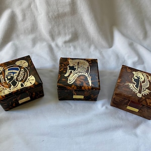 May include: Three small, square, wooden boxes with intricate designs. Each box features a different Egyptian-inspired image in gold on a brown background. The boxes are ideal for storing small trinkets or jewellery.
