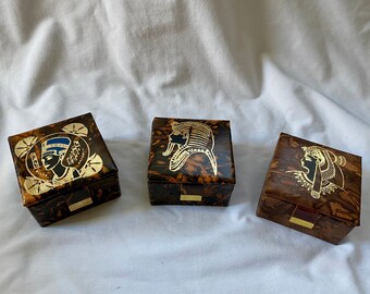May include: Three square boxes with a dark brown marbled finish and gold-colored metal latches. Each lid features a distinct gold-toned Egyptian-themed graphic, including a profile of Nefertiti with blue accents and two different pharaonic figures.