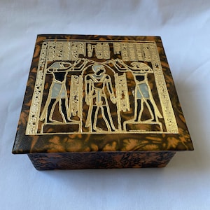 May include: A wooden box with a brown and orange patterned lid. The lid features a gold design of three Egyptian figures with hieroglyphics.
