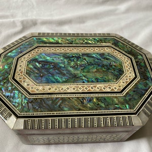 May include: An octagonal jewelry box with a shimmering abalone shell top. The box features intricate inlaid designs with black, white, and iridescent green and blue hues. The sides and edges are decorated with geometric patterns.