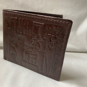 May include: A brown leather wallet with an embossed design of hieroglyphics. The wallet has a stitched edge and a flap closure.
