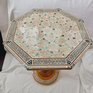 Peut inclure: Une table octogonale présentant des motifs de marqueterie géométrique complexes en bois clair, incrustés d'accents en nacre d'ormeau irisée. Le plateau affiche un design en mosaïque étoilée, tandis que le pied central est fini dans un ton brun chaud avec une bordure circulaire incrustée.